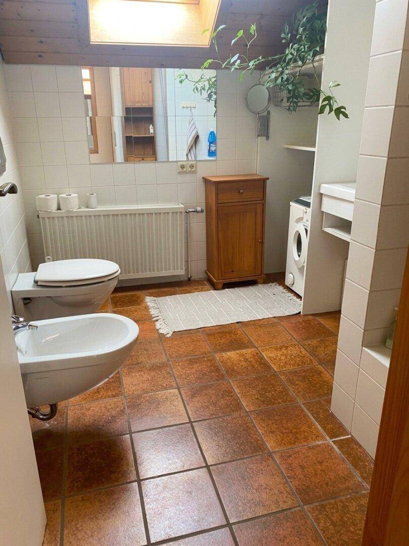 Badezimmer mit Terrakotta-Fliesen, Toilette, Bidet und Waschmaschine unter einem Dachfenster.