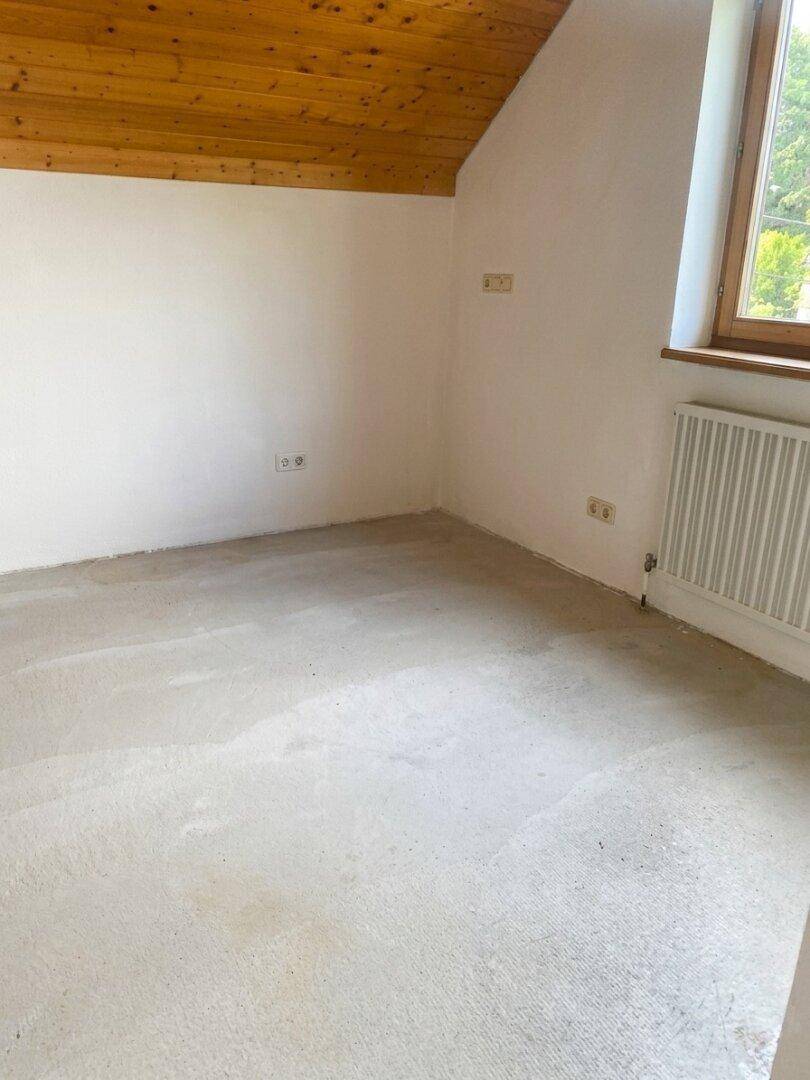 Leeres Zimmer mit weiß gestrichenen Wänden, Holzdachschräge und Betonboden.