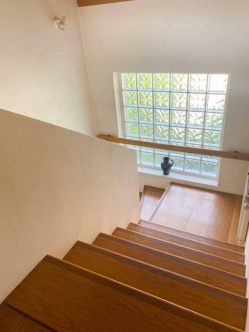 Holztreppe führt nach unten, beleuchtet durch ein großes Glasbausteinfenster.