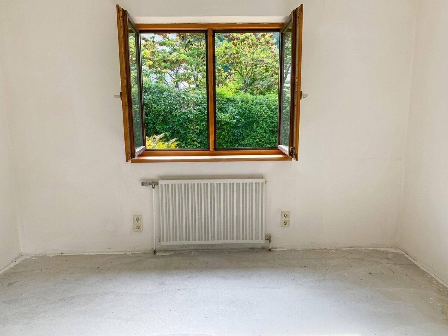 Helles Zimmer mit weiß gestrichenen Wänden, Fenster mit Blick ins Grüne und Heizkörper.