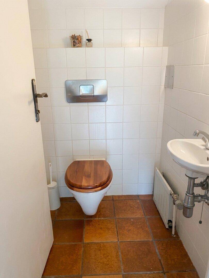 Kleines Gäste-WC mit weißen Fliesen, Terrakotta-Boden und einem WC mit Holzdeckel.