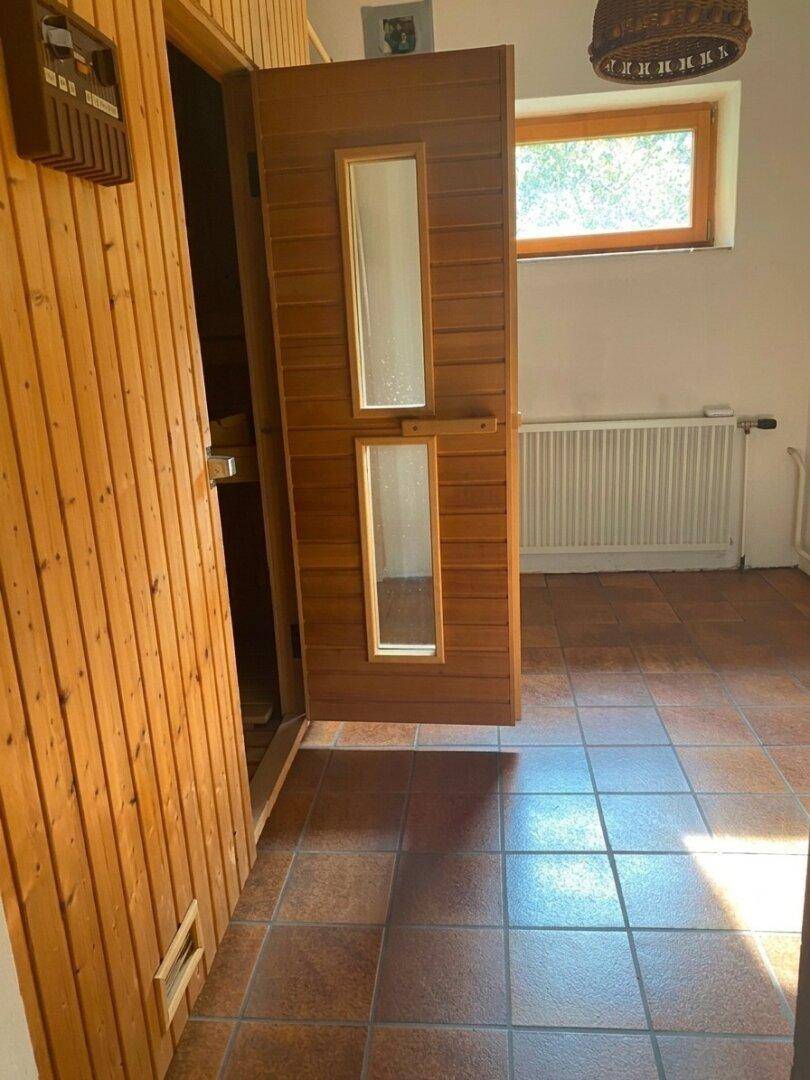 Eingang zu einer Holzsauna mit Terrakottafliesen und einem Fenster im Hintergrund.