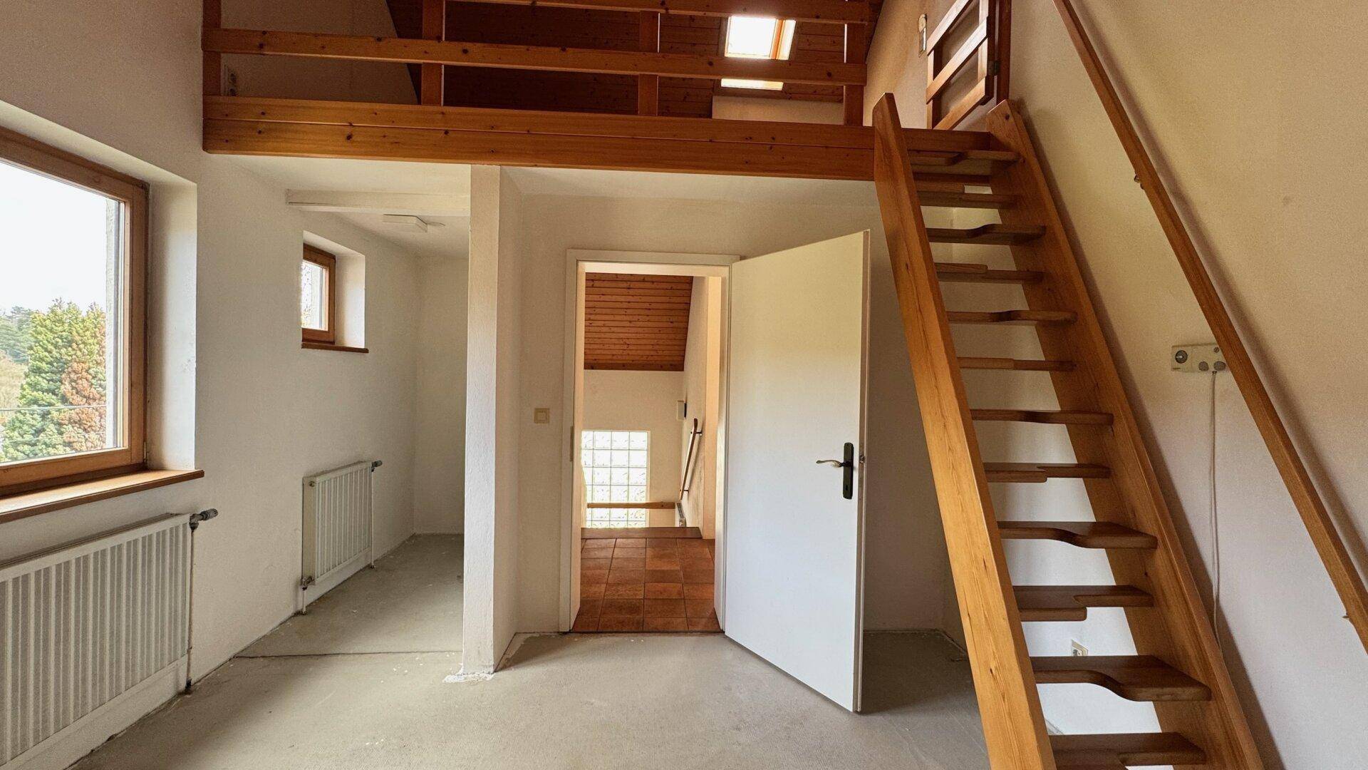 Heller Raum mit Betonboden, Holztreppe zu einer Galerie und Zugang zu weiteren Zimmern.