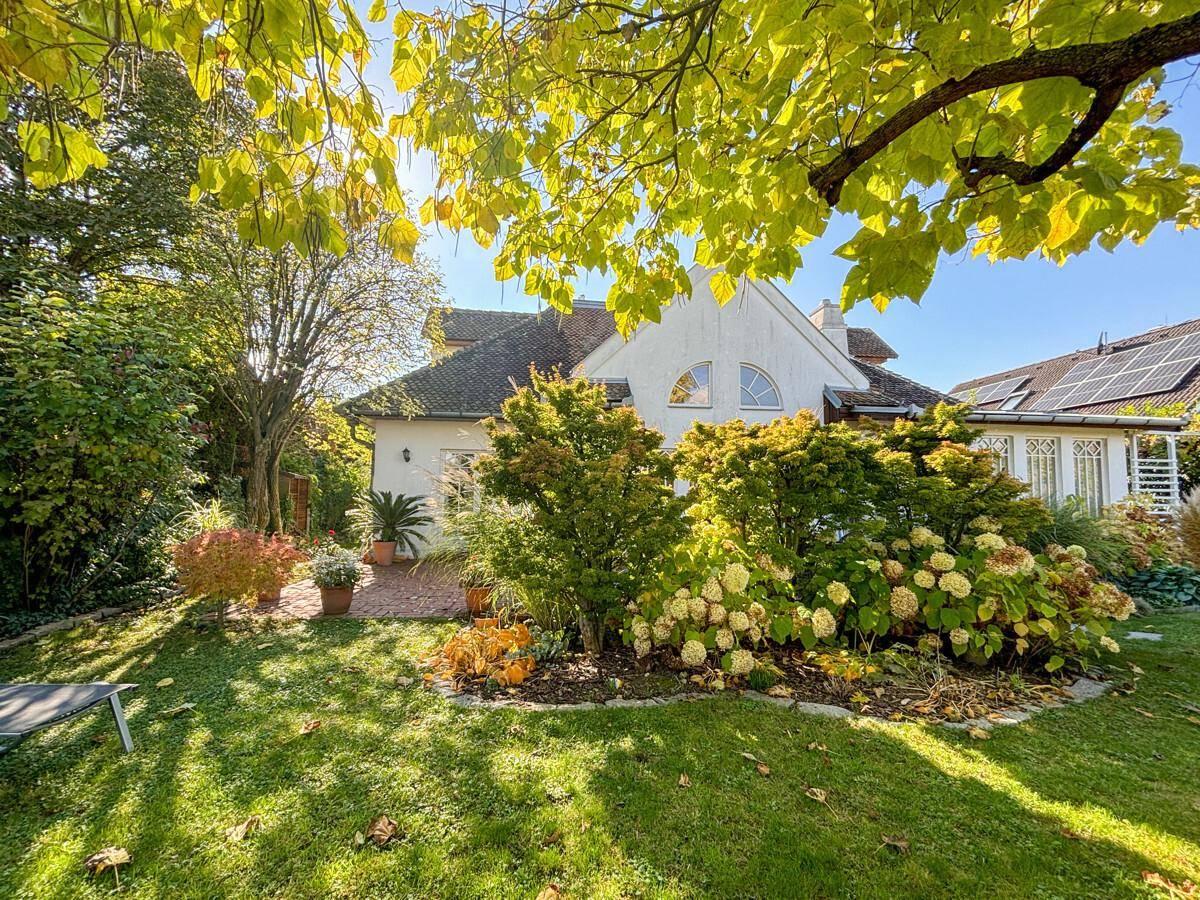 Sonnige Rückseite des Hauses mit üppigem Garten, grünen Bäumen und blühenden Sträuchern.