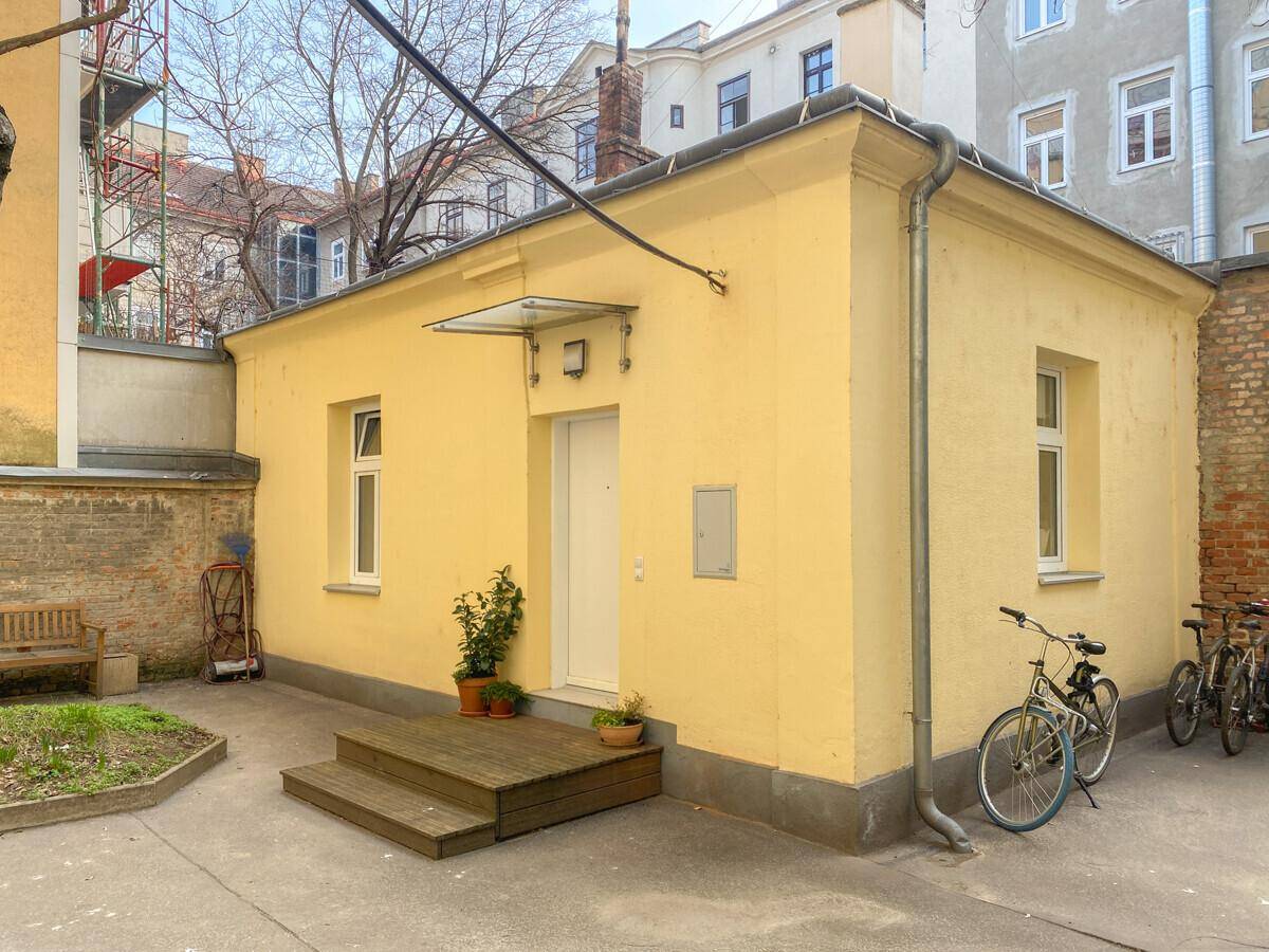 Gelbes Nebengebäude mit Eingangstür, Fenstern und einem kleinen Vorplatz mit Fahrrädern.