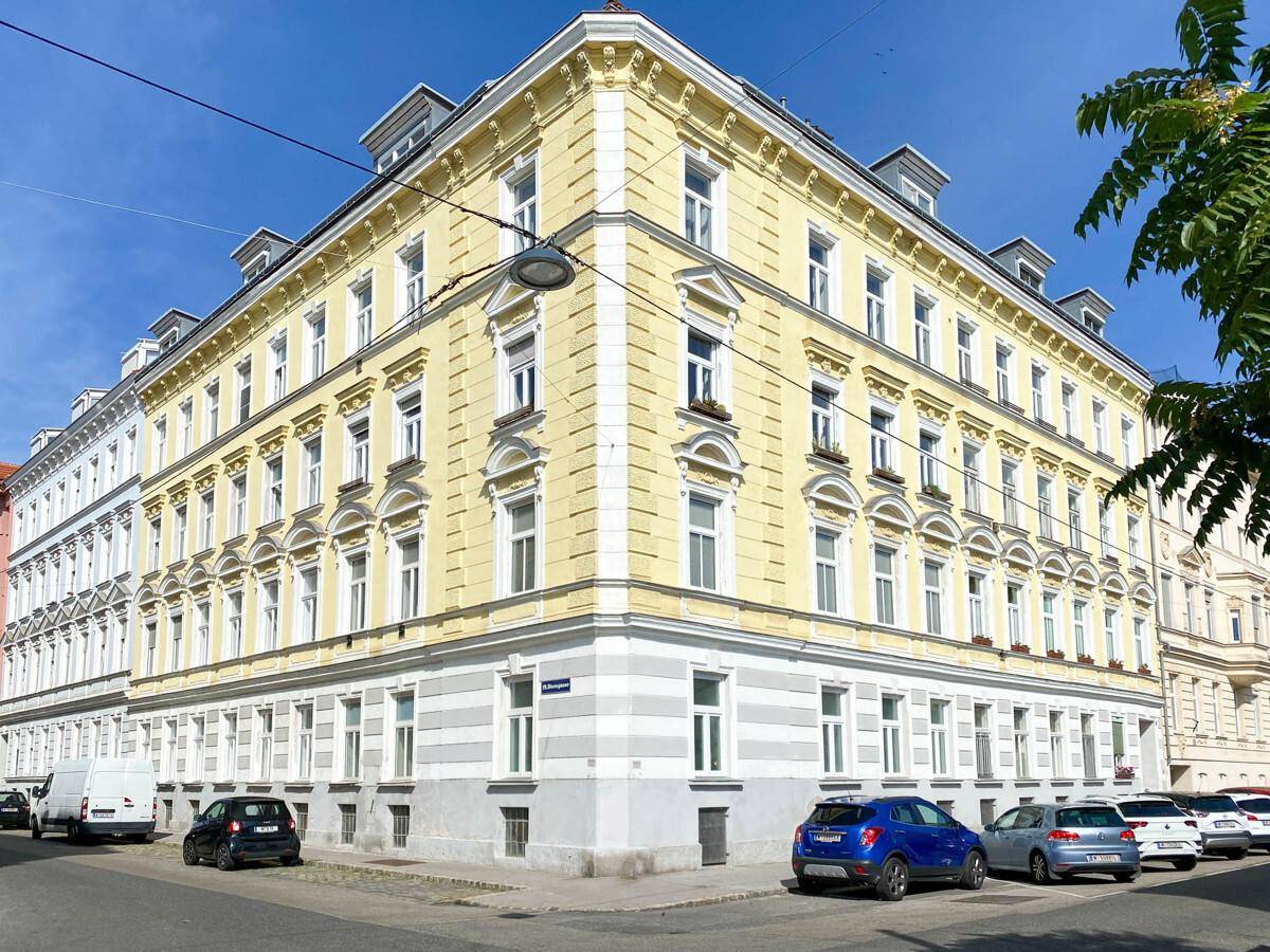 Eckgebäude mit klassischer Architektur und gelber Fassade in gutem Zustand, umgeben von städtischer Umgebung.