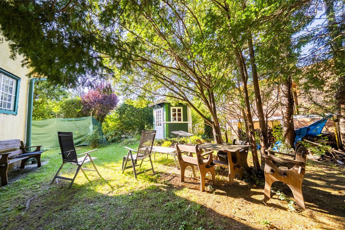 Gemütlicher Gartenbereich mit Sitzgelegenheiten im Schatten großer Bäume und einem kleinen Gartenhaus.
