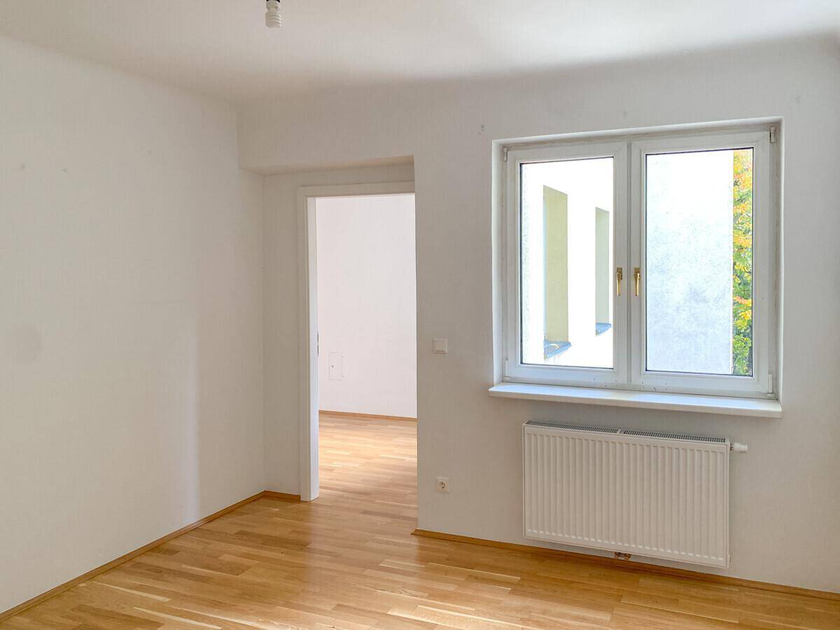 Helles Zimmer mit Parkettboden und offenem Durchgang zu einem weiteren Raum.