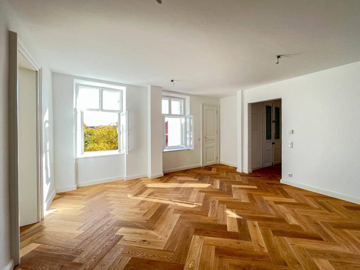 Geräumiges Zimmer mit hellem Fischgrätparkett und mehreren Fenstern, die den Raum erhellen.