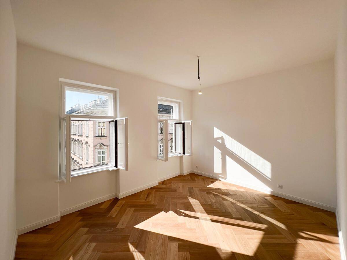 Helles Zimmer mit Fischgrätparkett und Blick aus den Fenstern auf die Stadtbebauung.