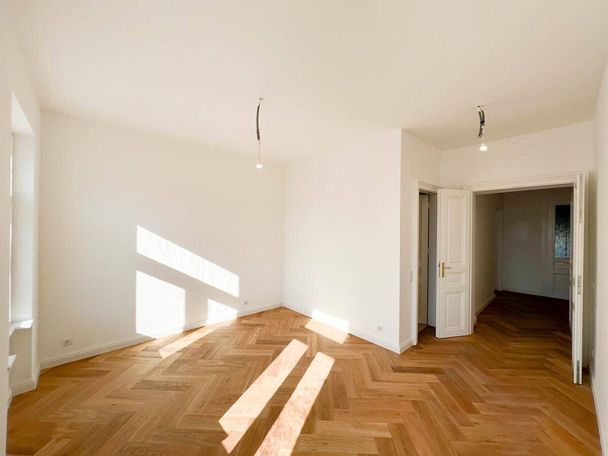 Lichtdurchflutetes Zimmer mit elegantem Fischgrätparkett und offener Tür zu einem weiteren Raum.