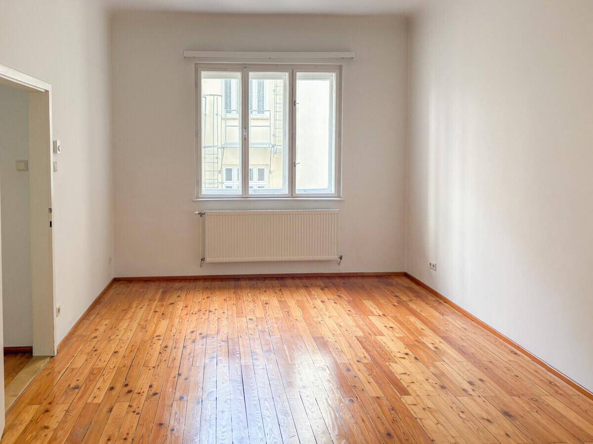 Geräumiges Zimmer mit glänzenden Holzdielen, weißen Wänden und einem großen Fenster.