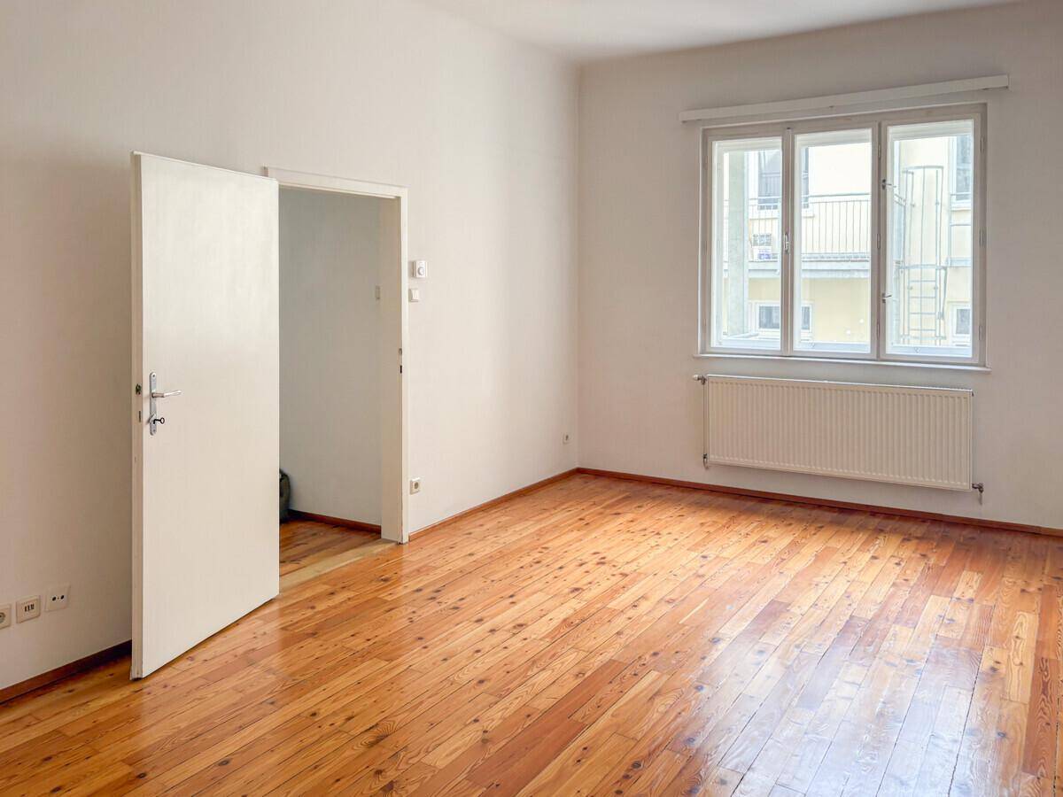 Helles, leeres Zimmer mit Holzdielenboden, weißen Wänden und einem Fenster zum Innenhof.