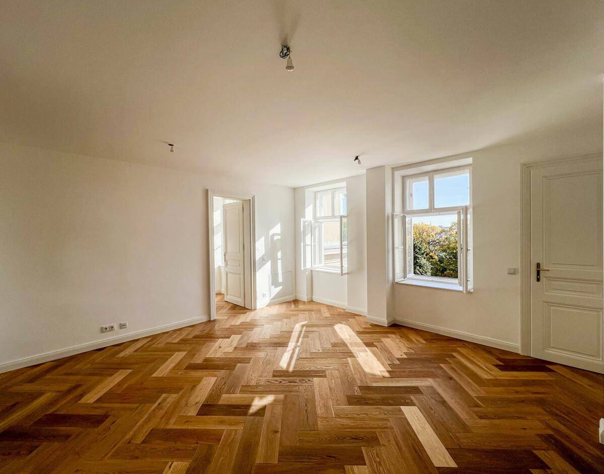 Helles, leeres Zimmer mit Fischgrätparkett und zwei Fenstern, die viel Sonnenlicht hereinlassen.