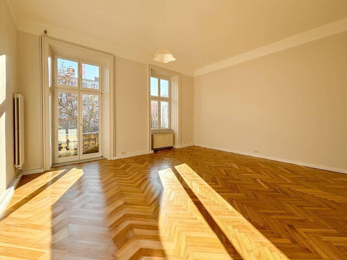 Helles, leeres Zimmer mit Fischgrätparkettboden und großen Fenstern, die viel Sonnenlicht hereinlassen.
