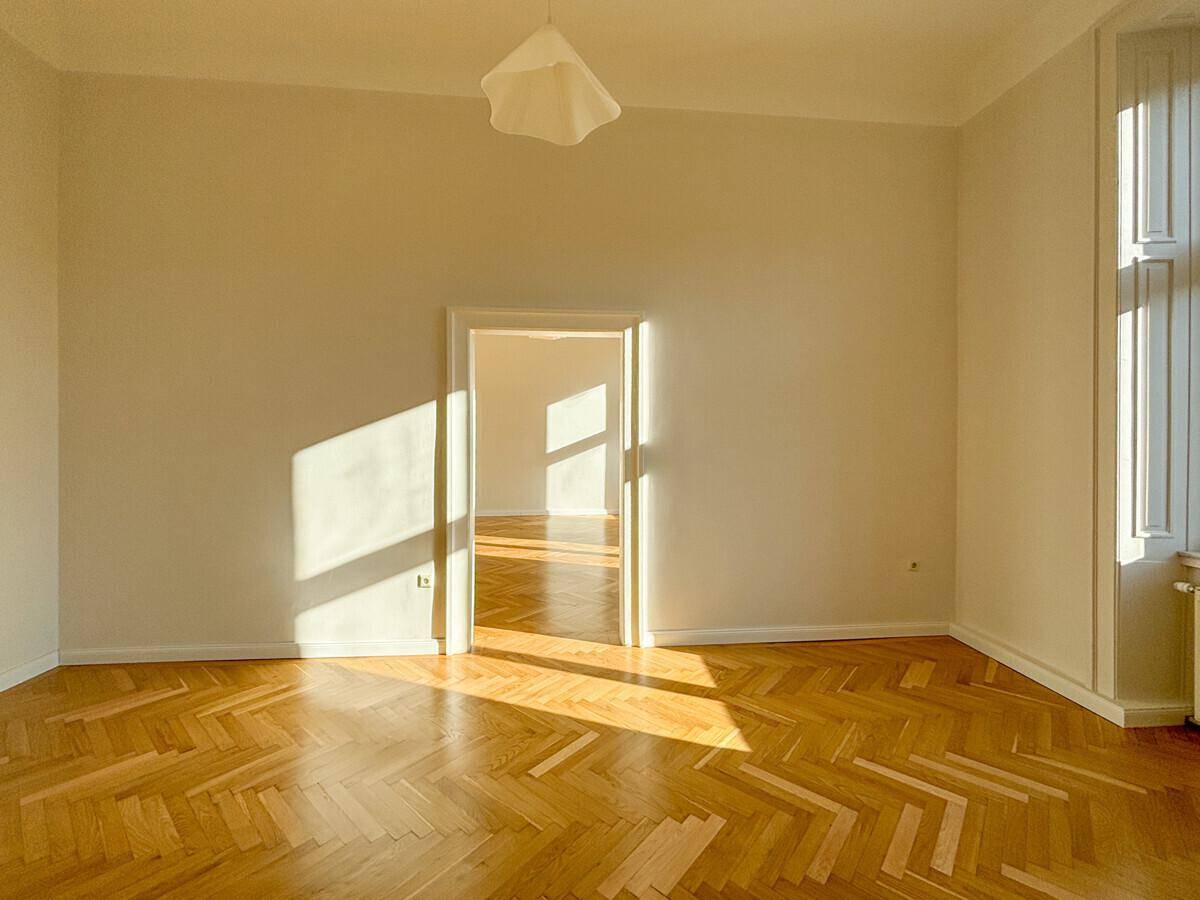 Leerer Raum mit Fischgrätparkett und offener Tür, die den Blick in einen weiteren sonnigen Raum freigibt.