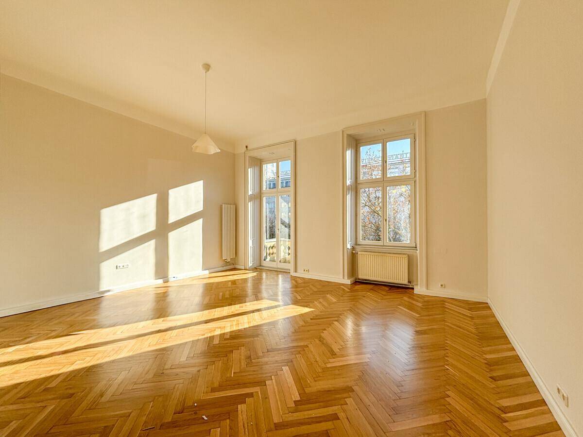 Großes, leeres Zimmer mit Fischgrätparkettboden und hohen Fenstern, die den Raum hell erleuchten.