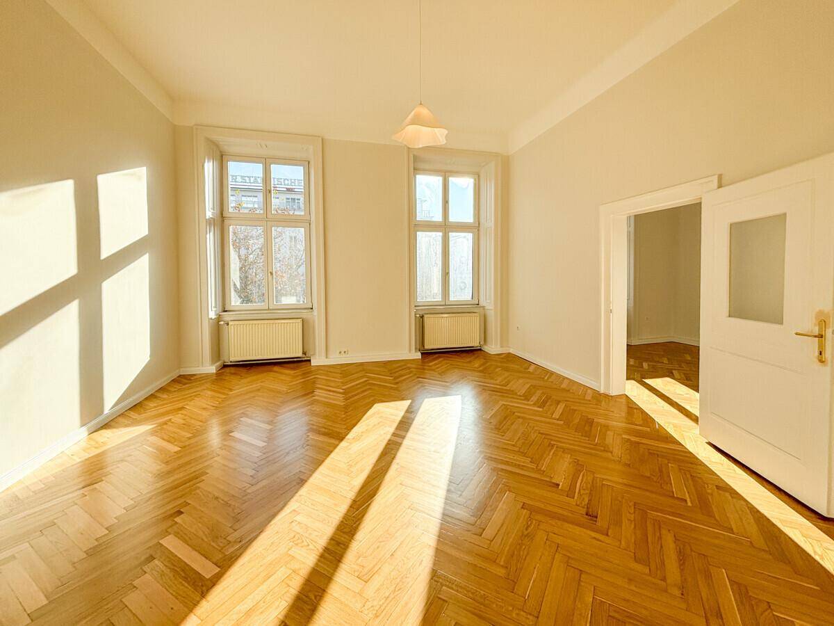 Geräumiges Zimmer mit Fischgrätparkett und Zugang zu einem weiteren Raum, hell durch große Fenster.