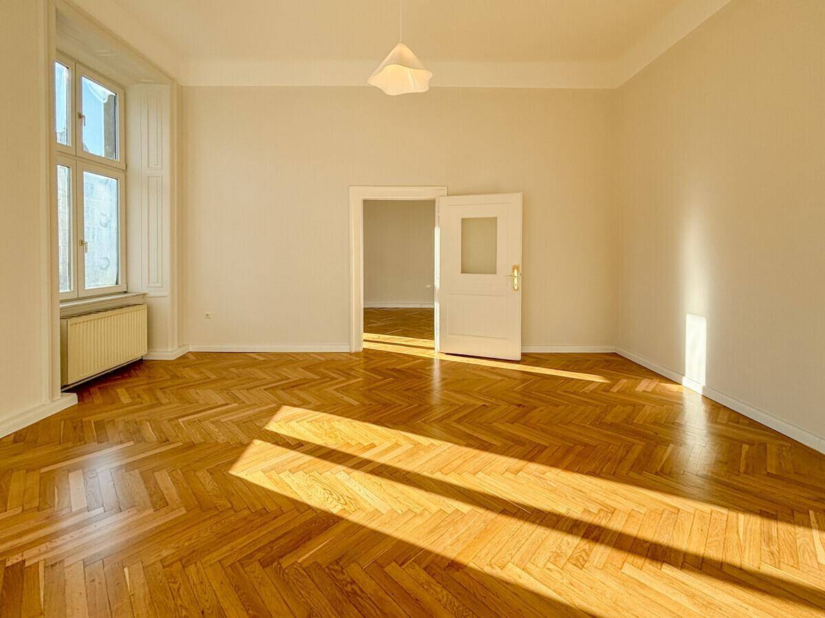 Heller Raum mit Fischgrätparkett und einem Fenster, das Sonnenlicht hereinlässt, mit offener Tür zu einem Nebenraum.