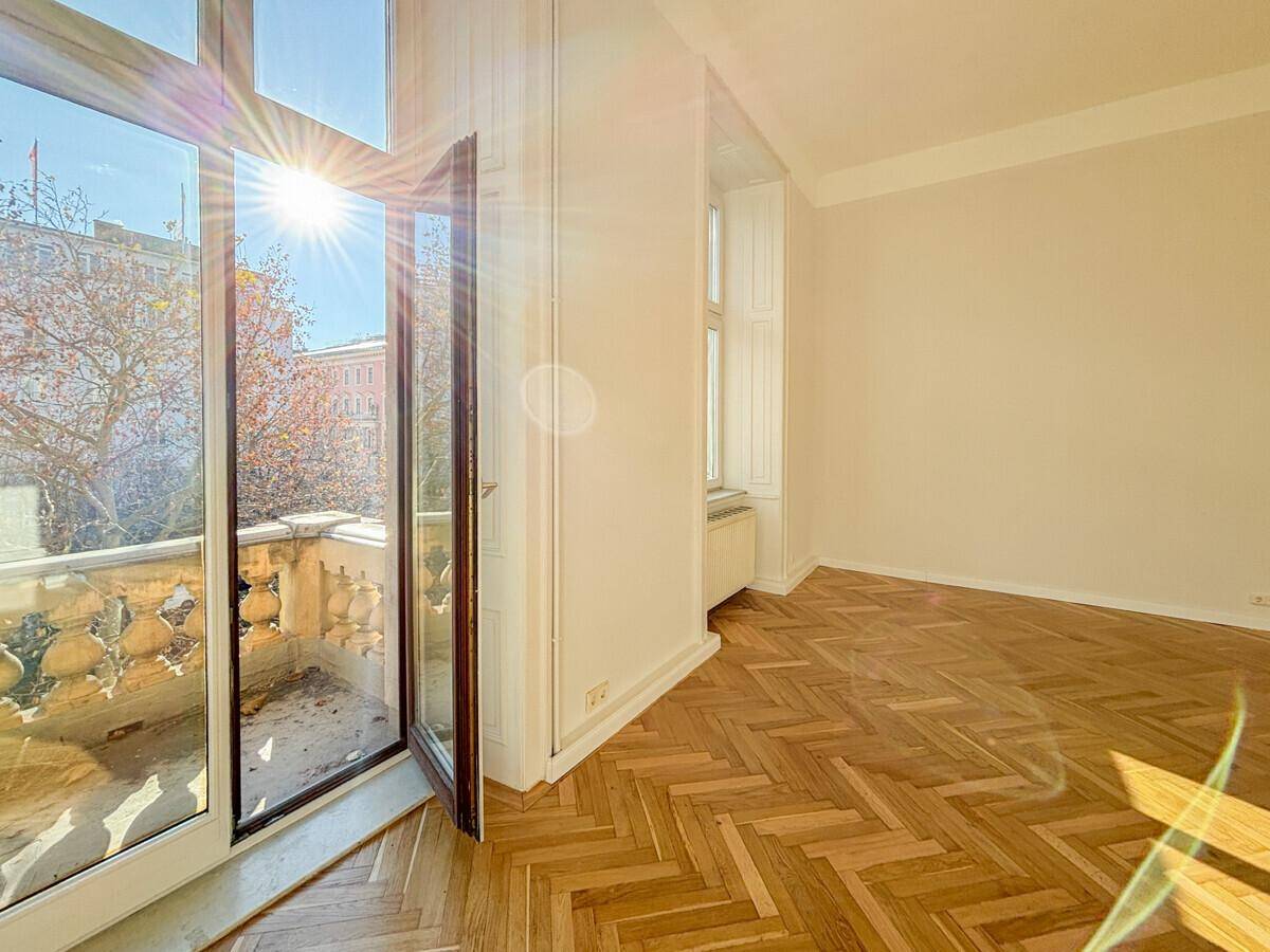 Lichtdurchflutetes Zimmer mit Fischgrätparkett und offenem Zugang zum Balkon, der Sonnenlicht hereinlässt.