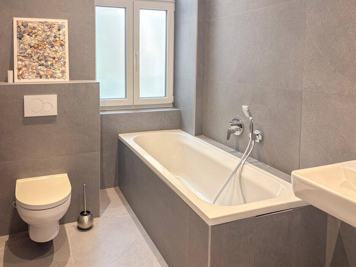 Modernes Badezimmer mit grauen Fliesen, integrierter Badewanne und wandhängendem WC.