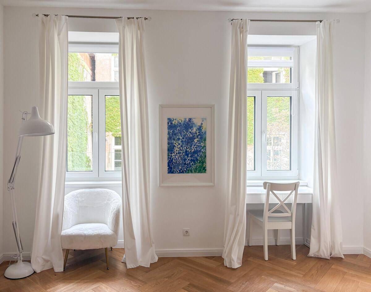 Heller Wohnbereich mit Schreibtisch am Fenster, weißem Sessel und Fischgrätparkettboden.