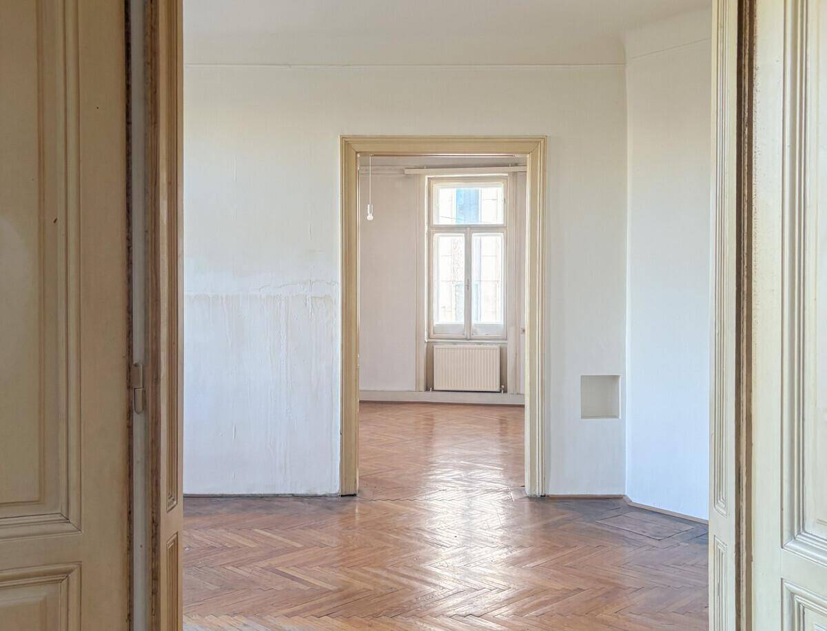 Heller Raum mit Fischgrätparkett, hohen Decken und Durchgang zu einem weiteren Zimmer.