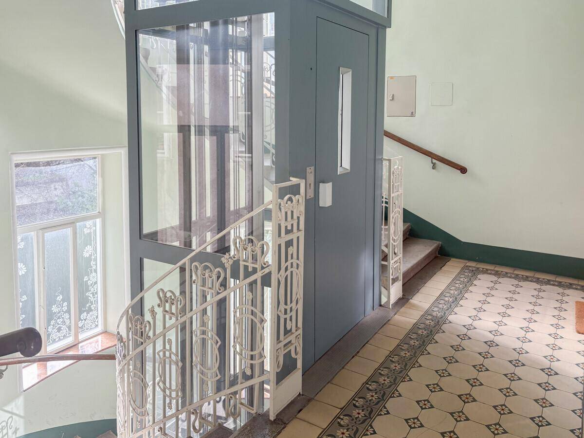 Moderner Glasaufzug und kunstvoll verziertes Treppengeländer in einem historischen Treppenhaus.