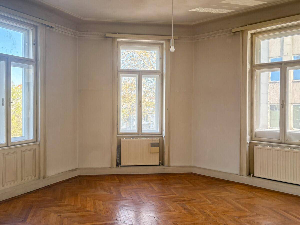 Helles Eckzimmer mit großen Fenstern, Fischgrätparkett und Stuckelementen an der Decke.