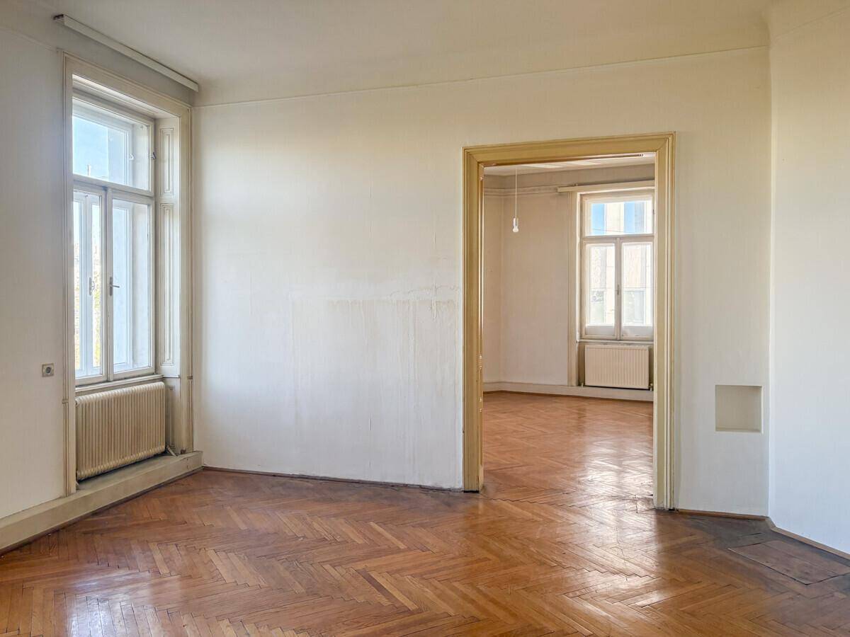 Geräumiges Zimmer mit Fischgrätparkett, hohen Fenstern und Blick in einen weiteren Raum.