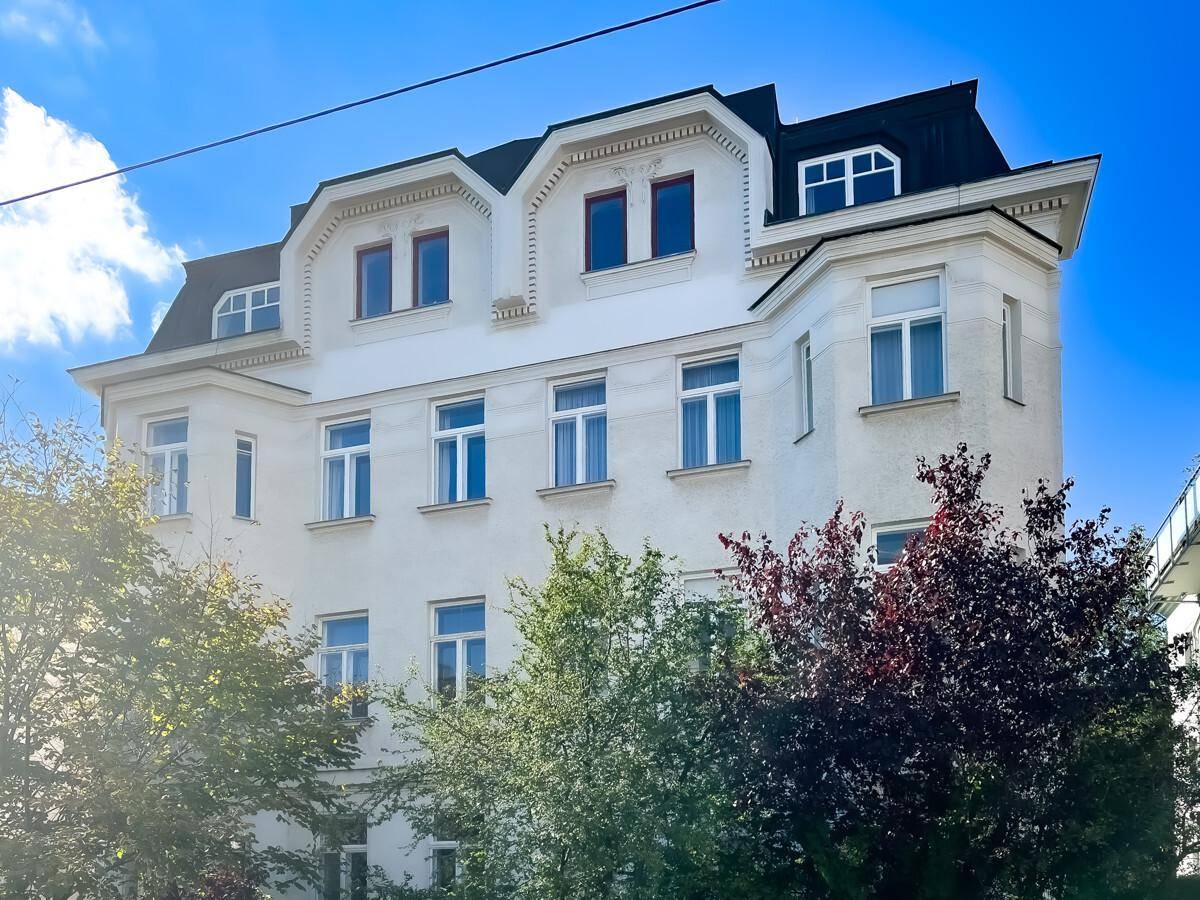 Historisches Gebäude mit weißer Fassade, vielen Fenstern und Mansarddach, umgeben von Bäumen.