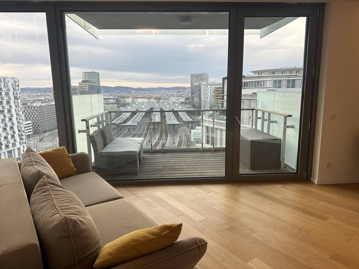 Helles Wohnzimmer mit beigem Sofa, Holzboden und Zugang zu einem Balkon mit Stadtblick.