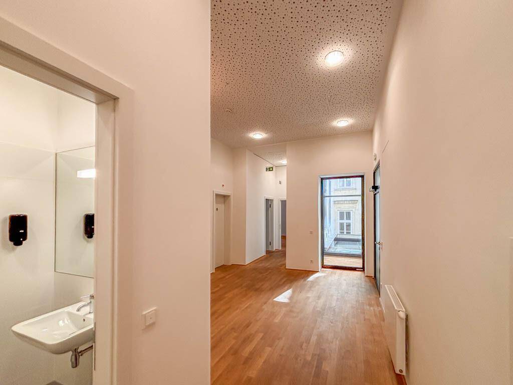 Heller Büroraum mit Holzboden und Zugang zu einem modernen Waschraum mit Waschbecken und Spiegel.