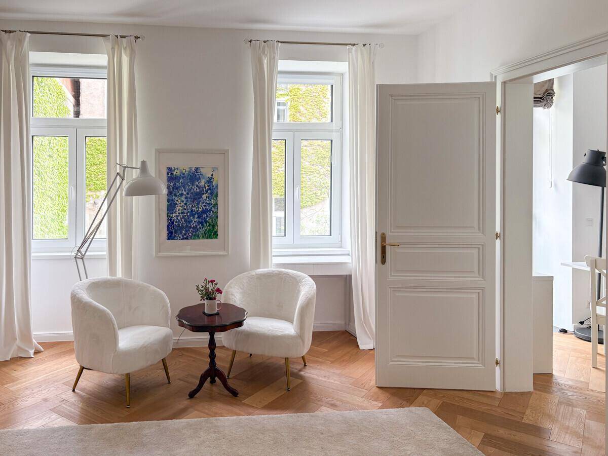 Heller Wohnbereich mit eleganten weißen Sesseln, Fischgrätparkett und großen Fenstern.
