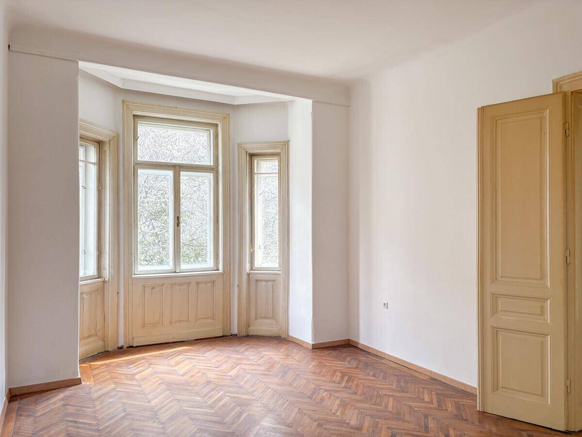 Geräumiges Zimmer mit Fischgrätparkett, großen Erkerfenstern und einer Holztür.