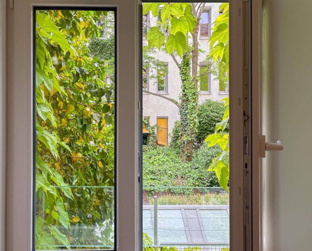 Blick aus einem Fenster in einen grünen Innenhof mit üppiger Vegetation und einem alten Gebäude.