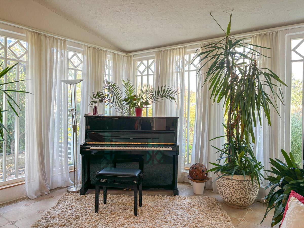 Heller Wintergarten mit vielen Fenstern, einem Klavier und großen Zimmerpflanzen.