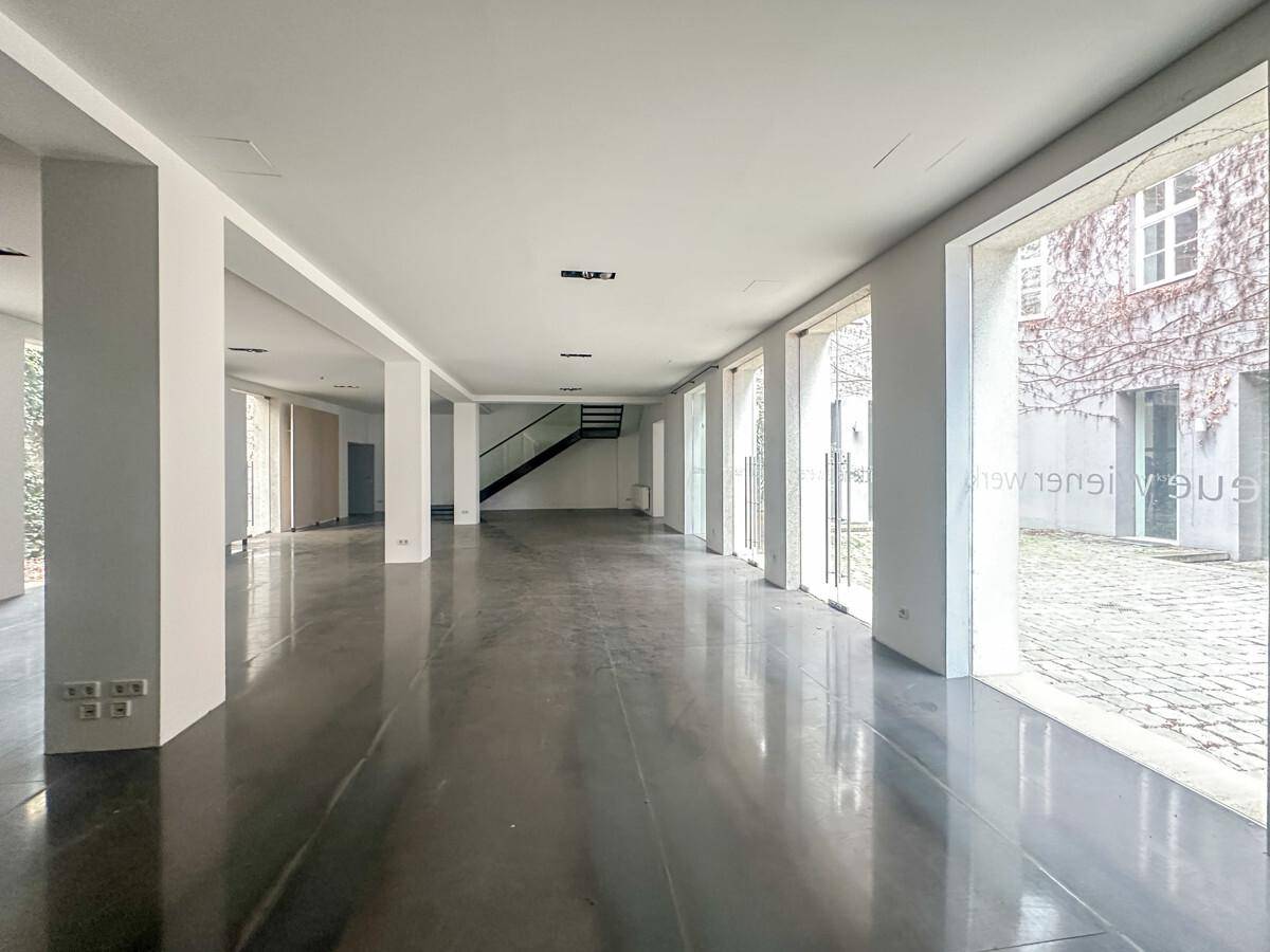 Großer, offener Raum mit Säulen, Betonboden und großen Fenstern, die zu einem begrünten Innenhof führen.