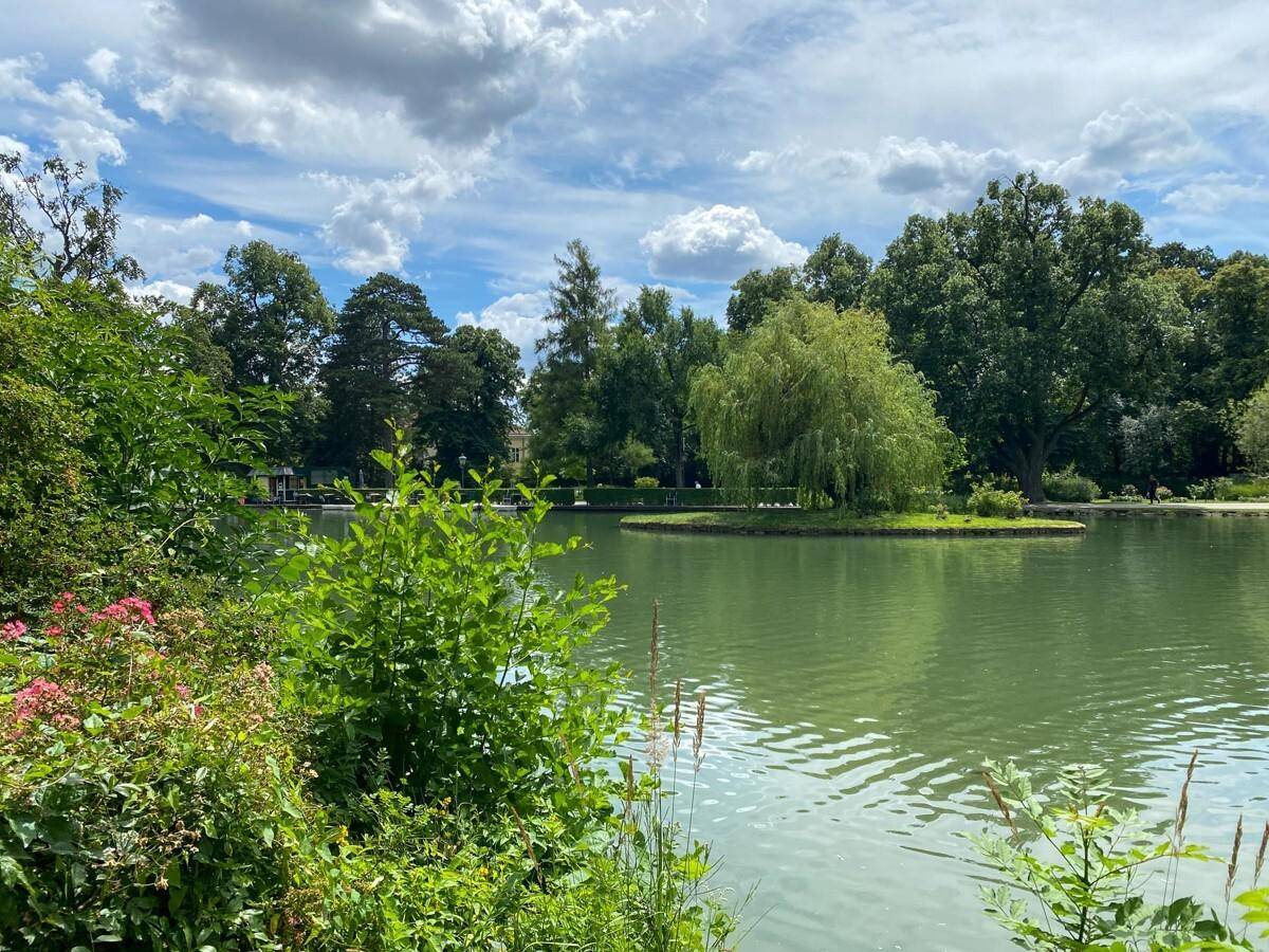 Ruhiger See mit einer kleinen Insel und üppiger Vegetation unter einem bewölkten Sommerhimmel.