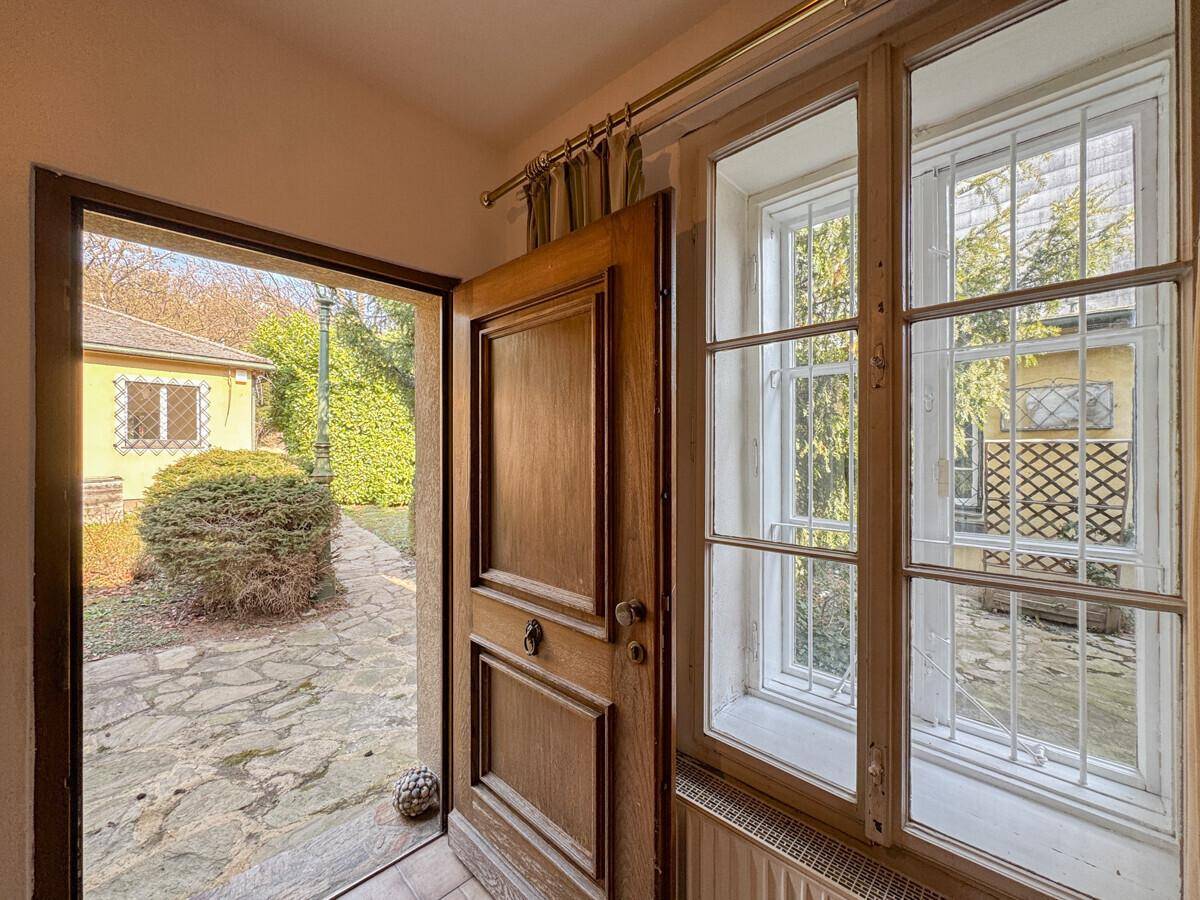 Eingangsbereich mit Holztür und Fenstern, die den Blick auf den gepflasterten Außenbereich freigeben.