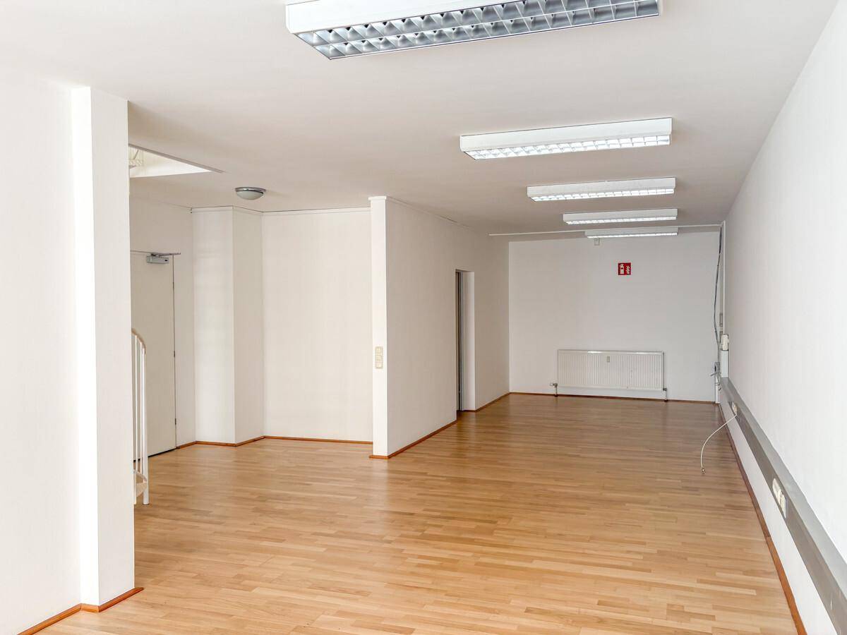 Langer, heller Büroraum mit Holzparkettboden, weißen Wänden und mehreren Deckenleuchten, ideal für offene Arbeitsbereiche.