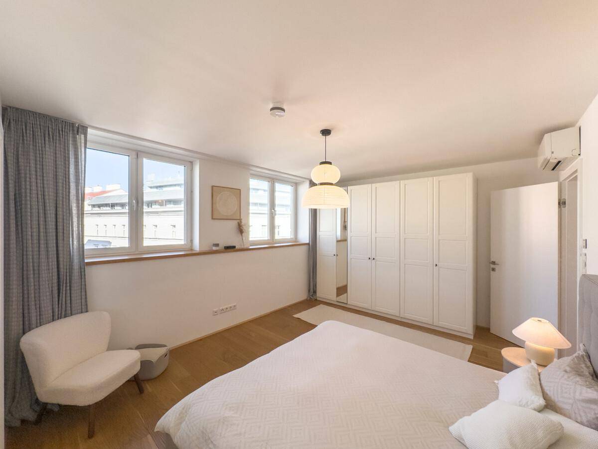 Helles Schlafzimmer mit weißem Kleiderschrank, Holzboden, großen Fenstern und Klimaanlage.