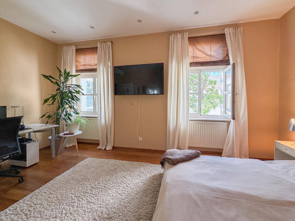 Helles Schlafzimmer mit Holzboden, großem Bett und Arbeitsbereich am Fenster.