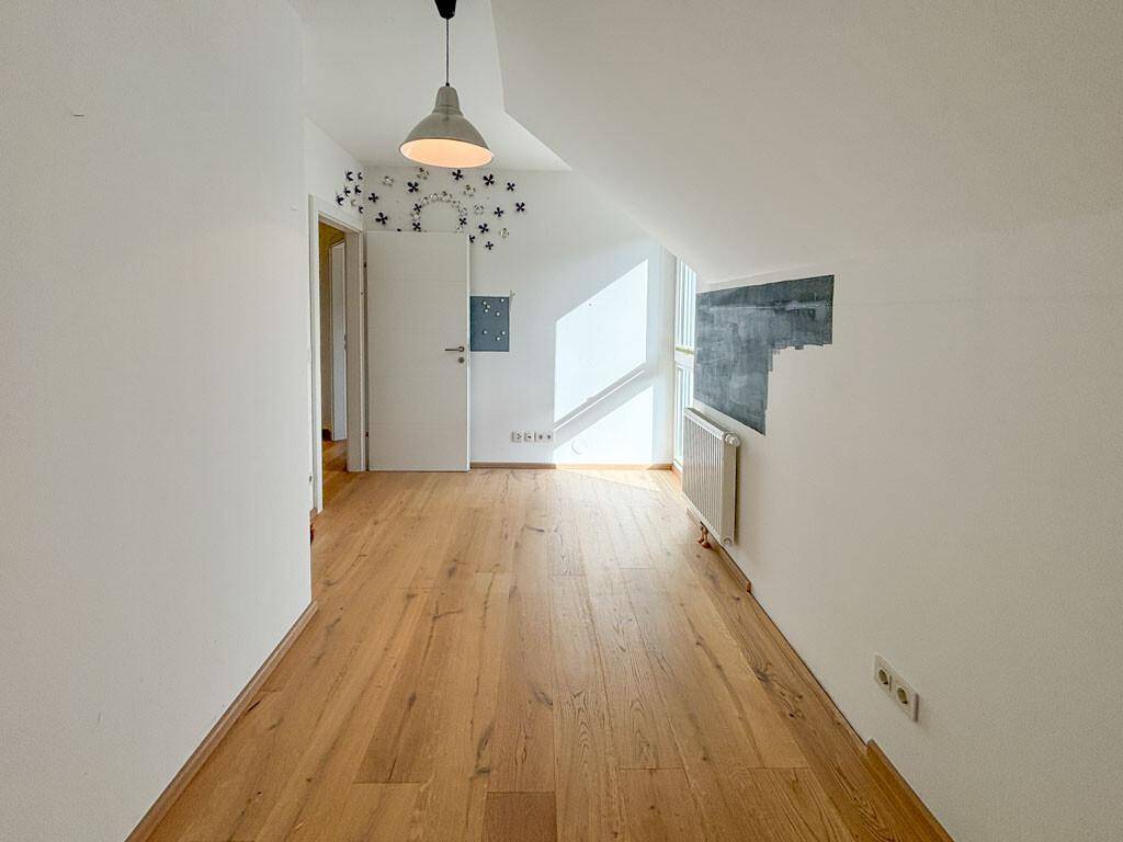 Leeres Zimmer mit Holzboden, viel Tageslicht und Zugang zu weiteren Räumen im Dachgeschoss.