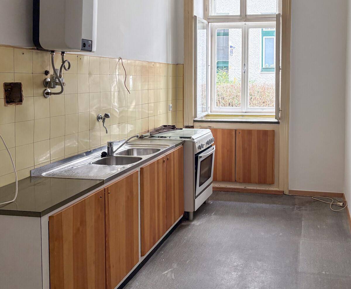 Küche mit Holzunterschränken, Doppelspüle, Herd und gefliestem Spritzschutz in Originalzustand.