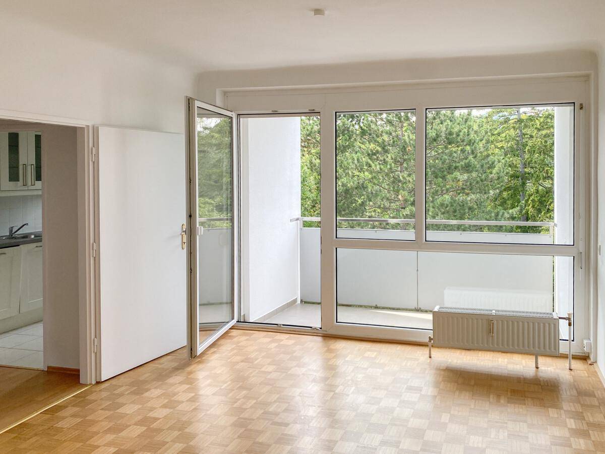 Helles Zimmer mit Parkettboden, großem Fenster und Zugang zum Balkon mit Waldblick.