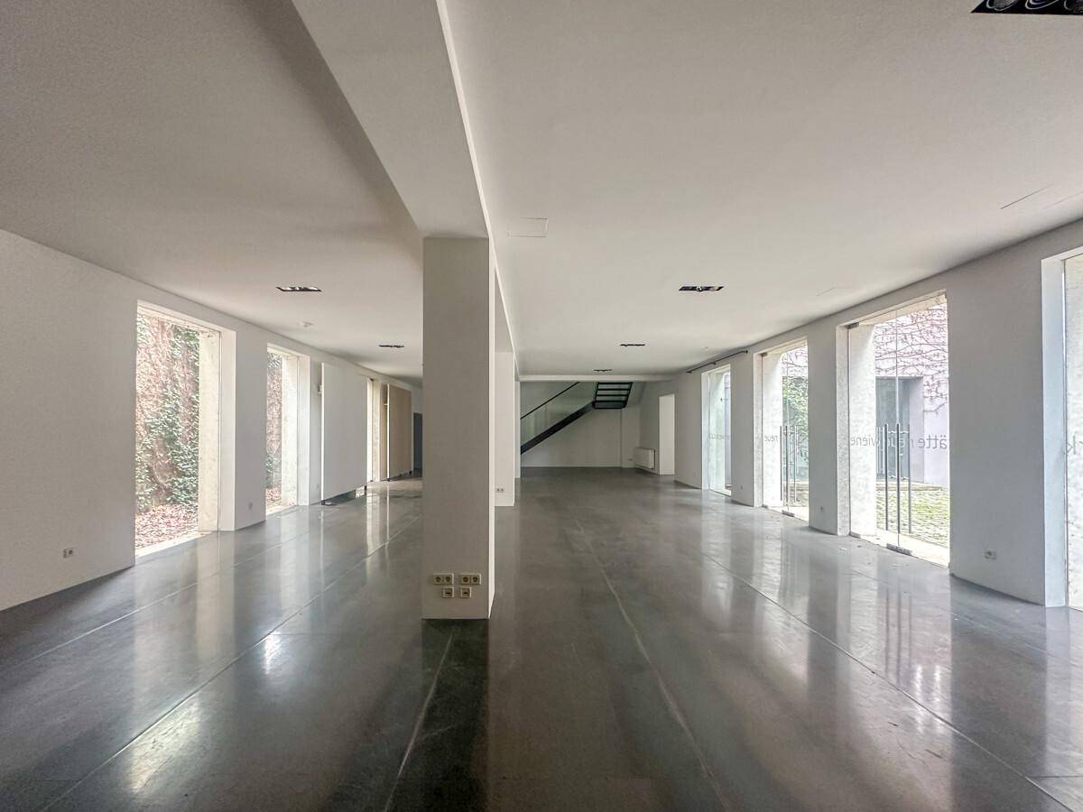 Weitläufiger, heller Raum mit Betonboden, Säulen und großen Fenstern, die einen Blick ins Grüne bieten.