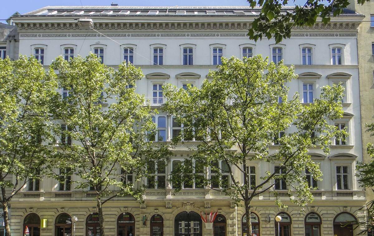 Prächtige historische Fassade eines Gebäudes mit vielen Fenstern und grünen Bäumen davor.