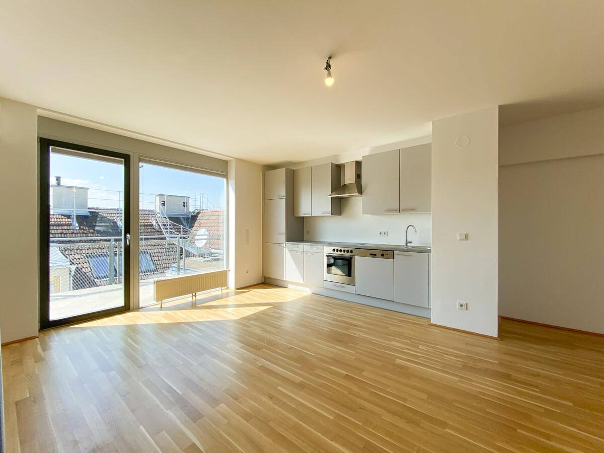 Heller offener Wohnbereich mit integrierter Küche, Parkettboden und Zugang zum Balkon.