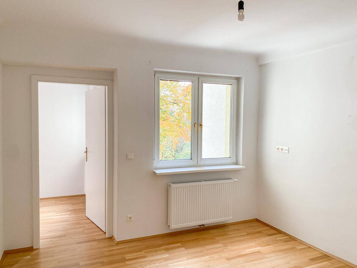 Helles Zimmer mit Parkettboden, weißen Wänden und einem Fenster, das viel Tageslicht hereinlässt.